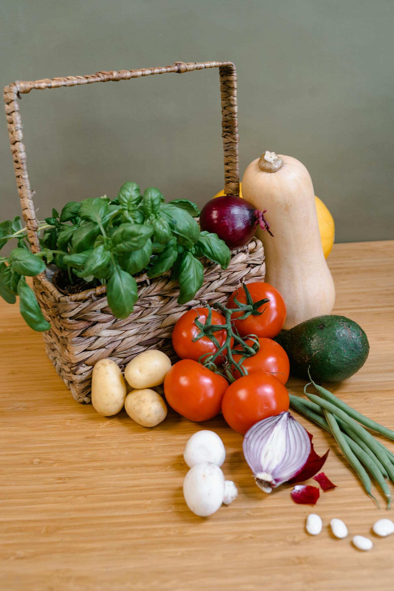 A vibrant assortment of fresh organic vegetables and basil leaves presented in a wicker basket on a wooden table.